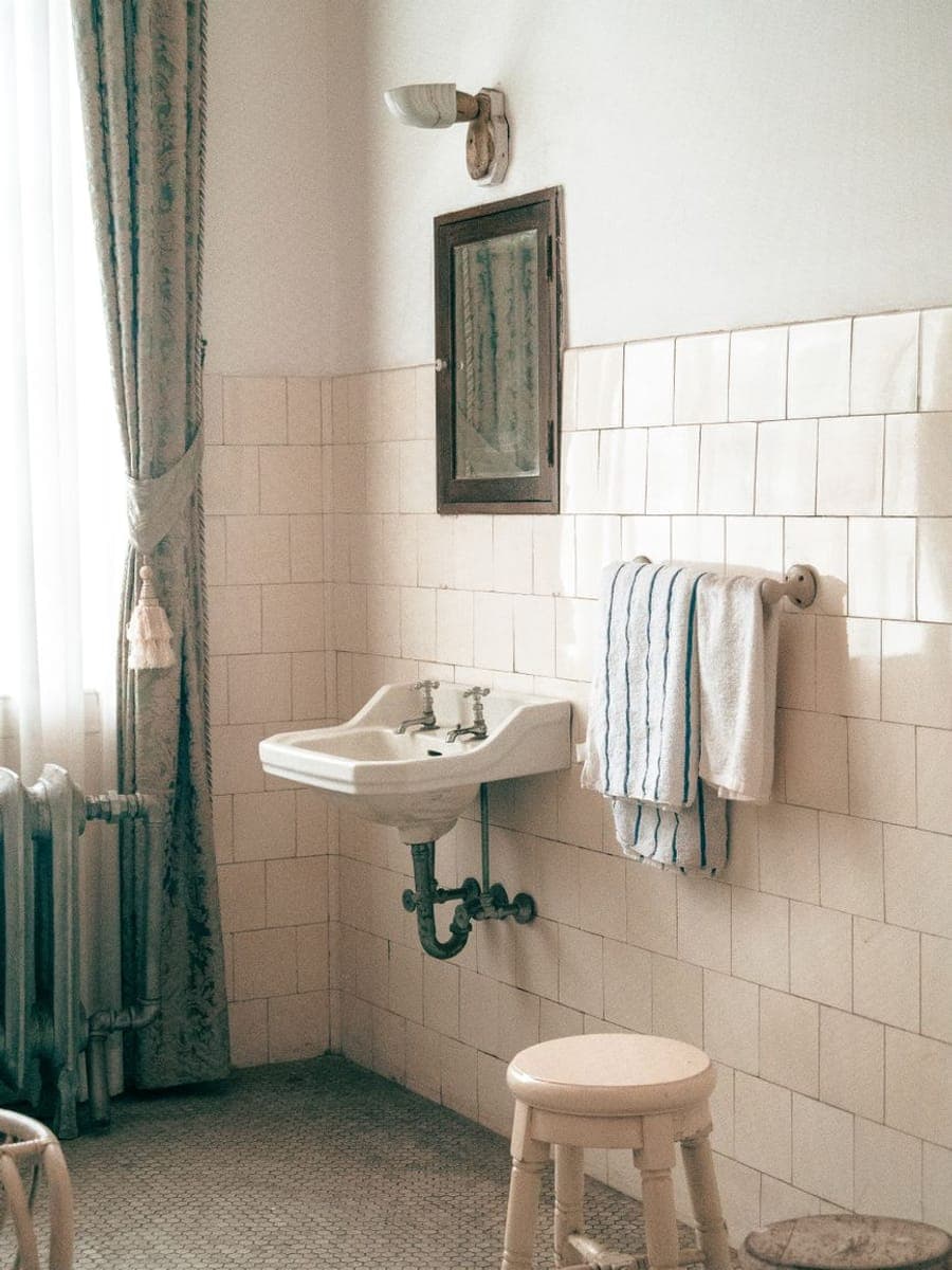 Baño antiguo con azulejo blanco a media altura, lavabo de porcelana con dos grifos, radiador de hierro y suelo de mosaico