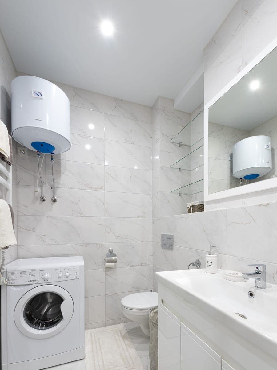 Baño estrecho de mármol blanco con un termo eléctrico horizontal en alto, el dial del termostato a la vista y la lavadora bajo él