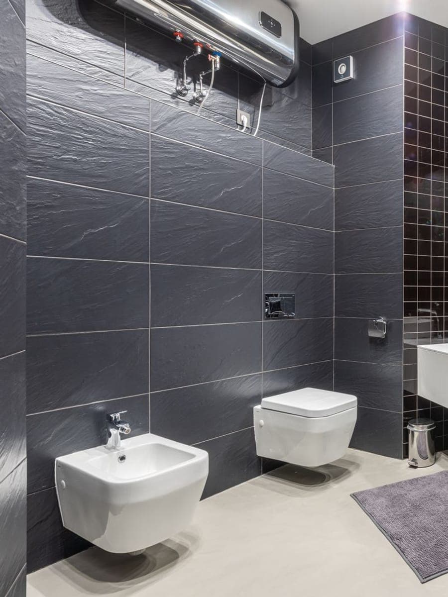 Baño de azulejo gris oscuro con un termo eléctrico horizontal colgado en lo alto de la pared, junto a la mampara de ducha y el lavabo suspendido