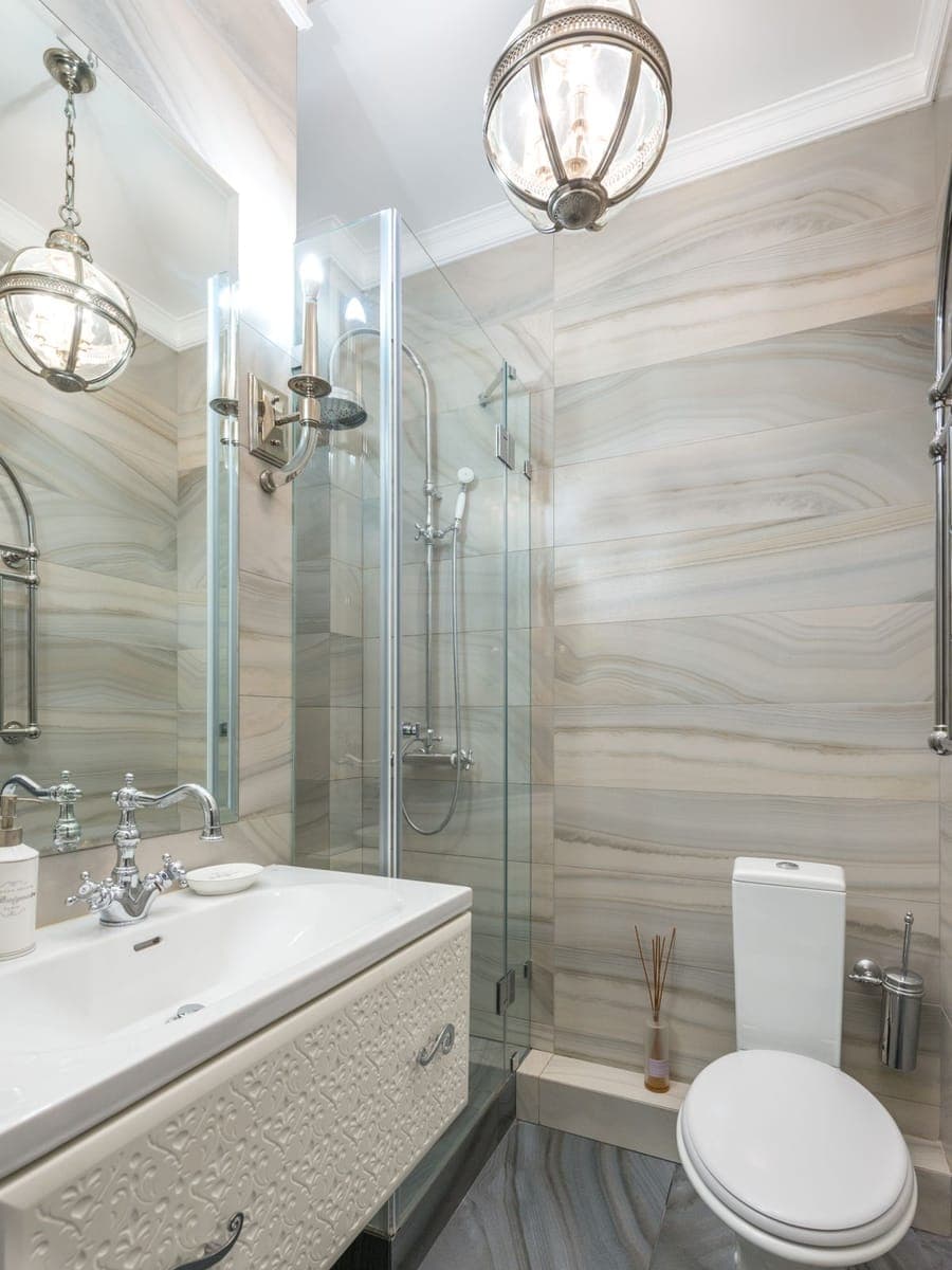 Baño alargado con paredes de azulejo veteado, lavabo sobre mueble blanco, mampara de ducha de cristal e inodoro al fondo