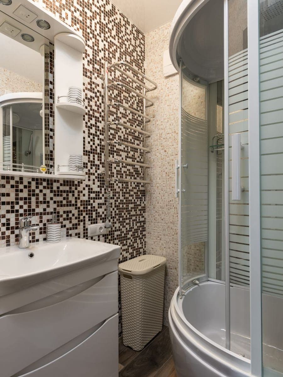 Baño estrecho de mosaico marrón con toallero radiador cromado, lavabo sobre mueble blanco y bañera con mampara curva