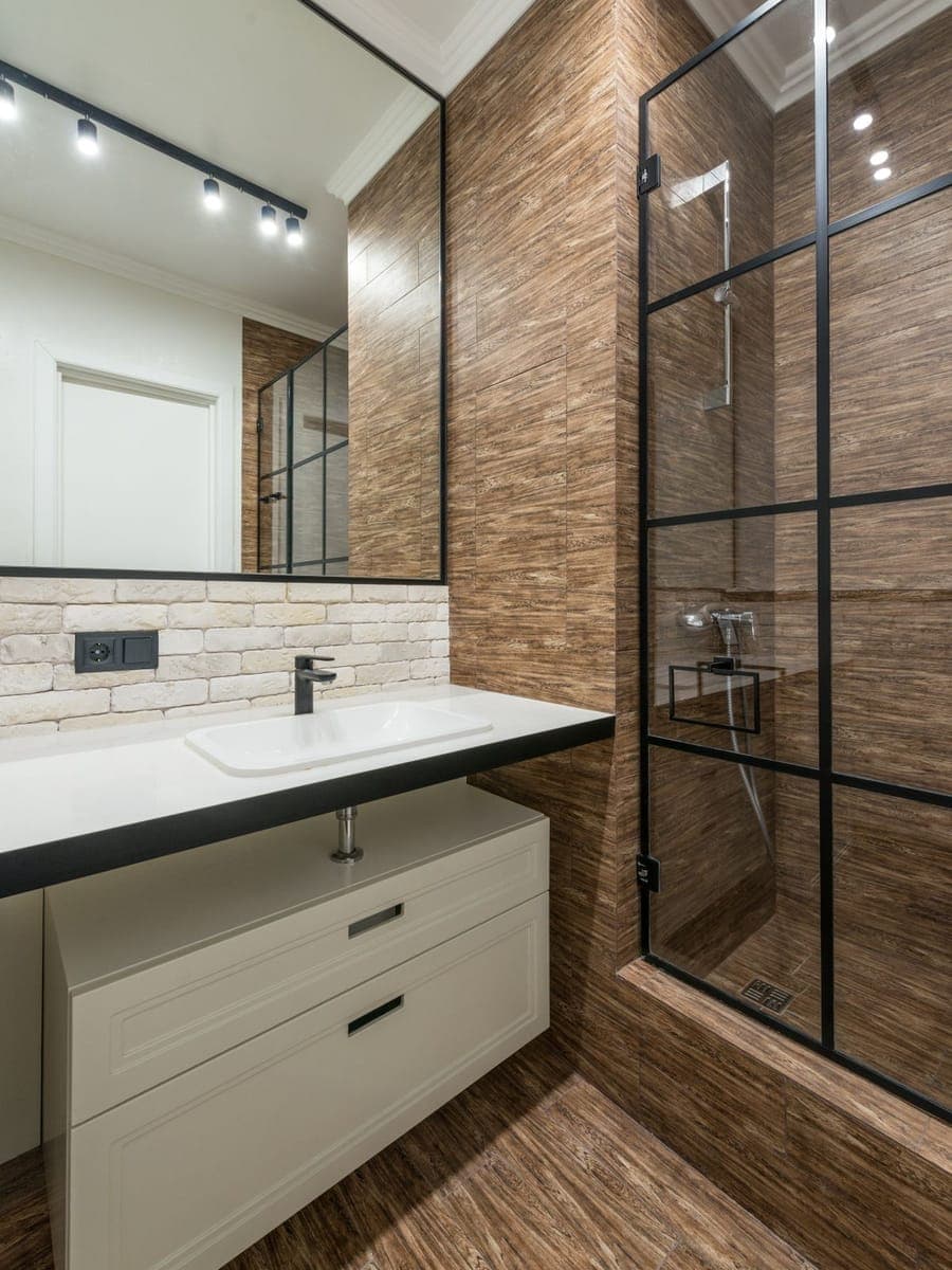 Baño con pared de azulejo imitación madera, lavabo sobre encimera oscura, zócalo de ladrillo visto con un enchufe y una mampara de ducha de perfil negro