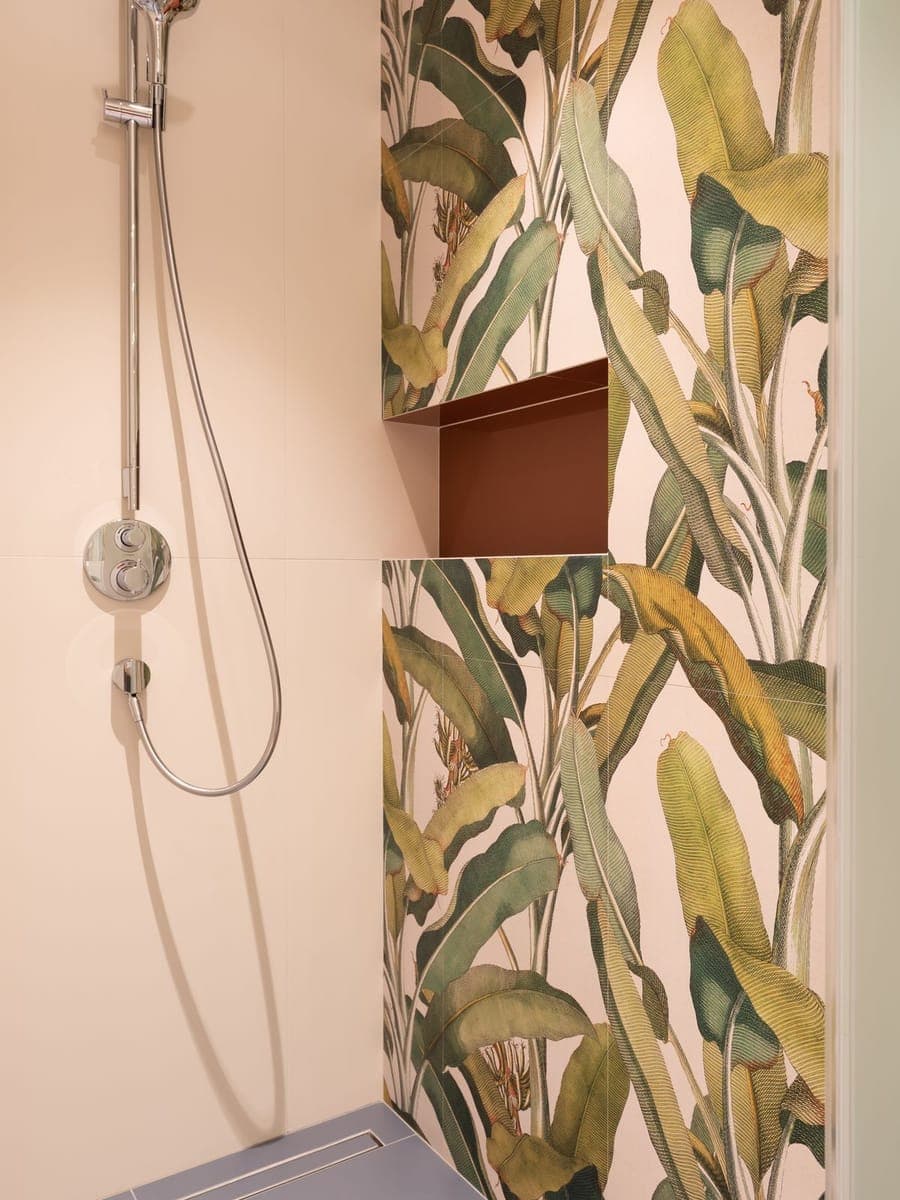 Rincón de ducha con grifo termostático cromado y flexible, junto a una pared alicatada con dibujo de hojas verdes y una hornacina