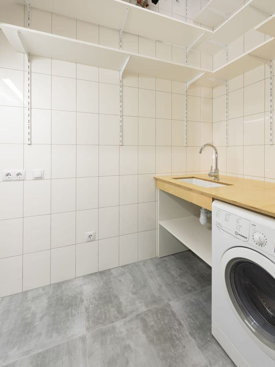 Cuarto de lavado alicatado en blanco con baldas metálicas, encimera de madera con fregadero y lavadora, y tubos con llaves en lo alto