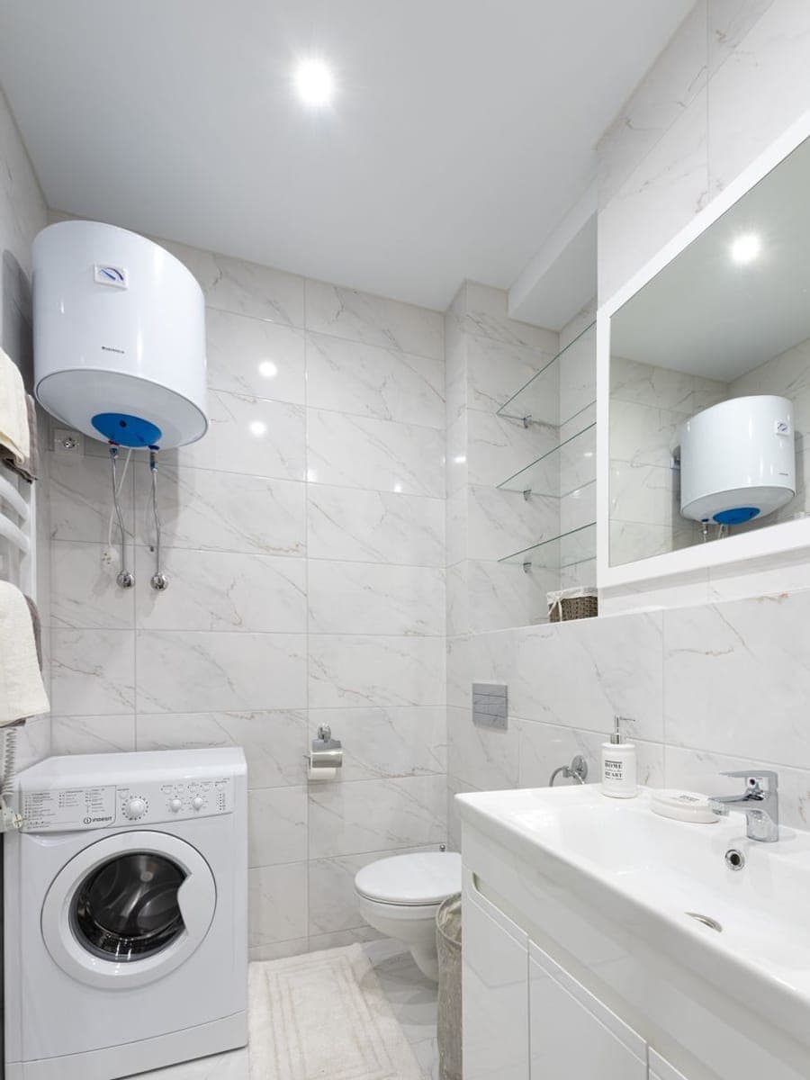 Termo eléctrico blanco colgado en alto en un baño de mármol claro, con los dos latiguillos bajando y la lavadora debajo