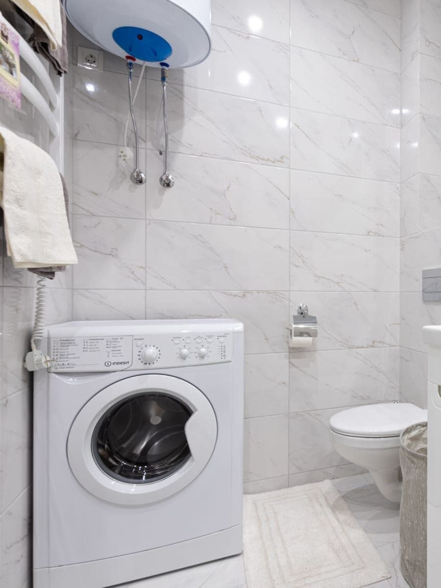Termo eléctrico blanco colgado en horizontal en lo alto de un baño de mármol claro, con los dos latiguillos flexibles bajando hasta la pared y una lavadora debajo