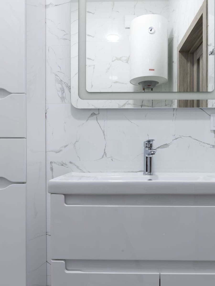 Lavabo blanco con grifo cromado bajo un espejo en el que se refleja un termo eléctrico vertical colgado en la pared de enfrente