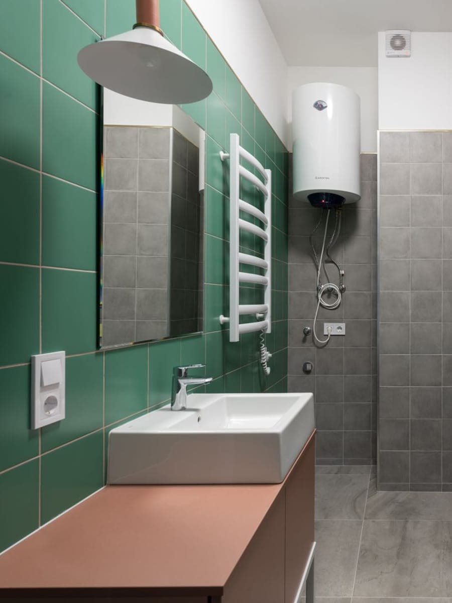 Termo eléctrico vertical blanco colgado en la pared de un baño de azulejo verde y gris, entre un toallero radiador y la bañera con mampara