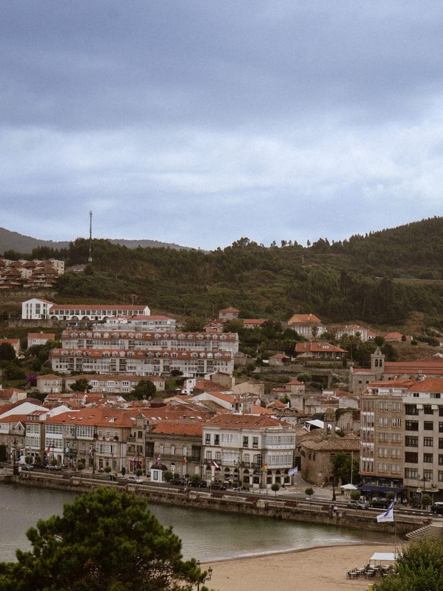 Villa costera de tejados rojos sobre una playa y una ría, con monte arbolado detrás y cielo nublado