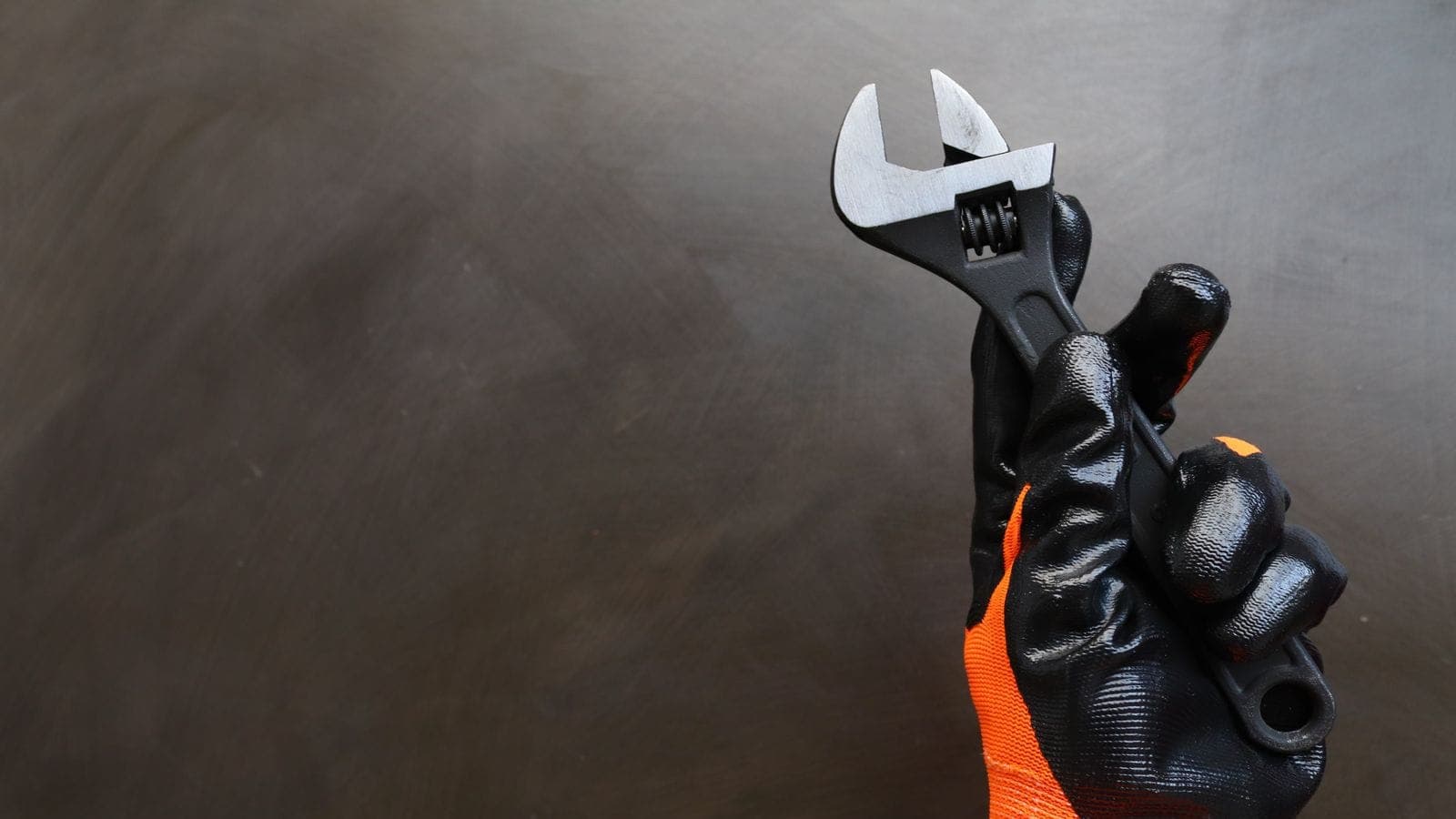 Mano con guante negro y naranja de trabajo sujetando una llave ajustable sobre un fondo oscuro