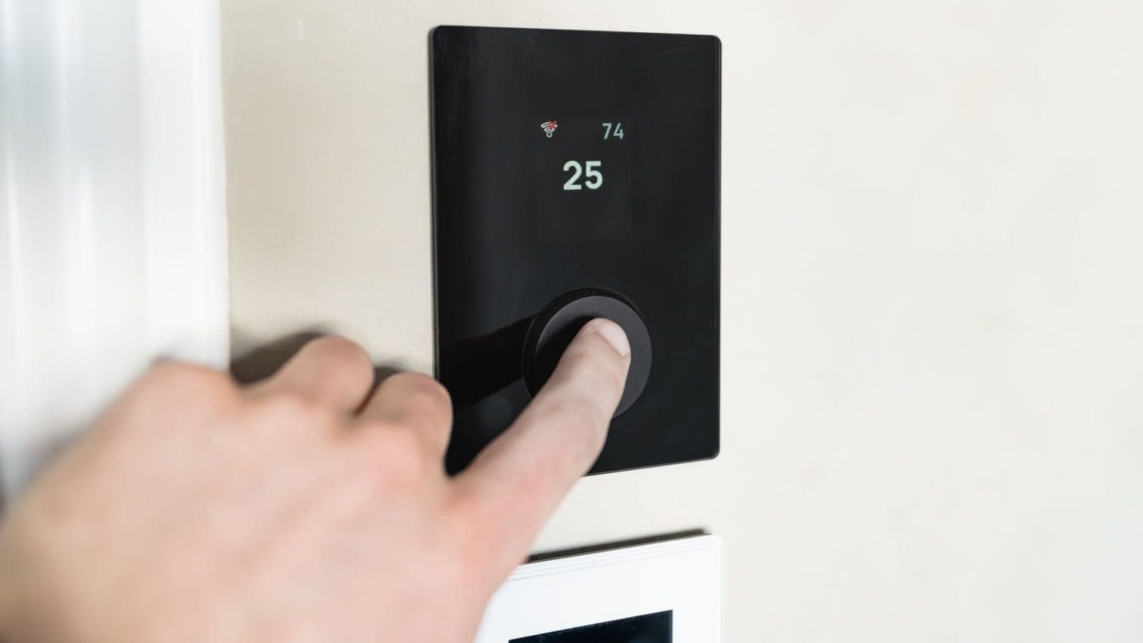 Un dedo pulsando la rueda de un termostato digital negro empotrado en la pared, con la pantalla marcando 25