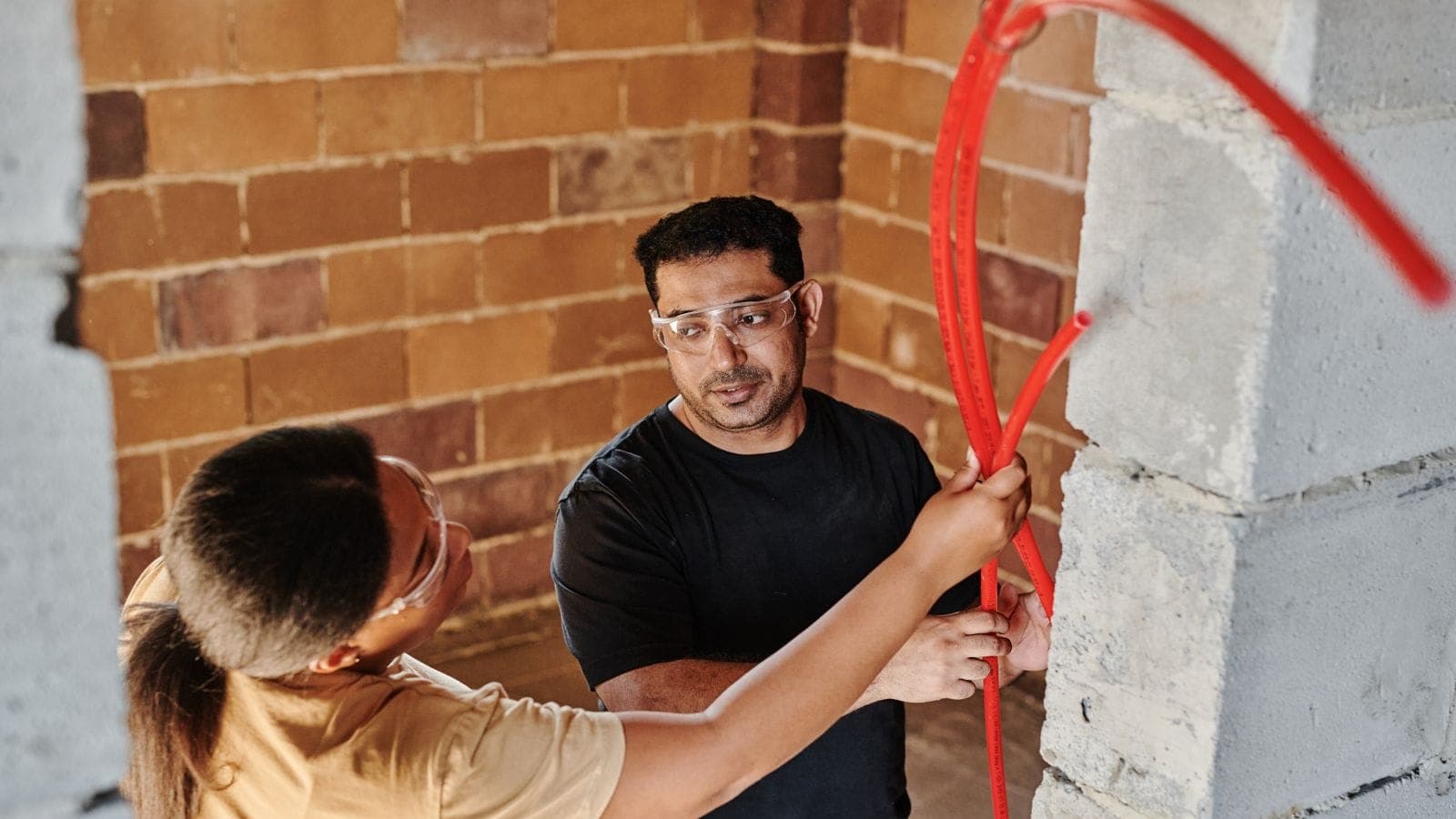 Dos operarios con gafas de protección pasando un tubo rojo de fontanería por una pared de bloque