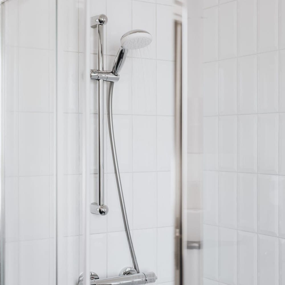 Barra de ducha cromada con teléfono y agua cayendo, en una ducha de azulejo blanco