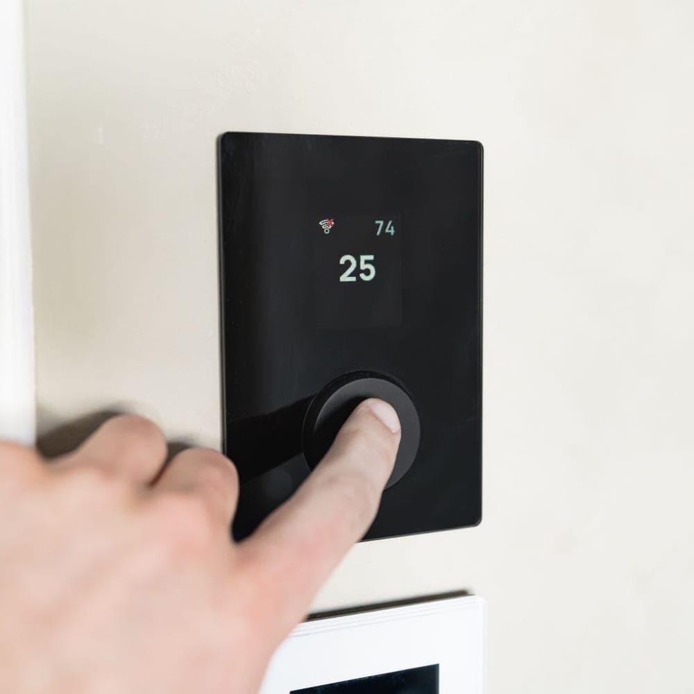 Un dedo pulsando la rueda de un termostato digital negro empotrado en la pared, con la pantalla marcando 25
