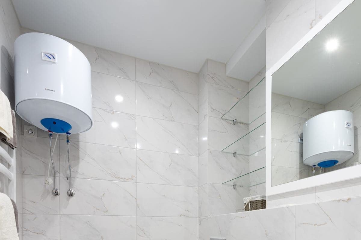 Baño estrecho de mármol blanco con un termo eléctrico horizontal en alto, el dial del termostato a la vista y la lavadora bajo él