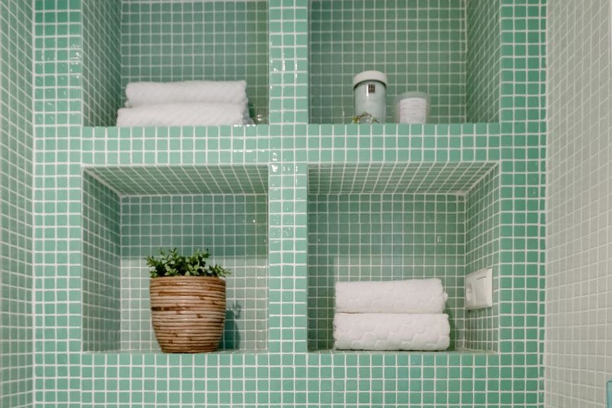 Baño de mosaico verde agua con dos hornacinas alicatadas, toallas dobladas y una planta, junto a un lavabo blanco