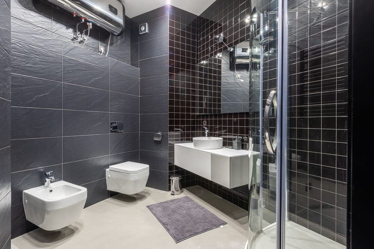 Baño de azulejo gris oscuro con un termo eléctrico horizontal colgado en lo alto de la pared, junto a la mampara de ducha y el lavabo suspendido
