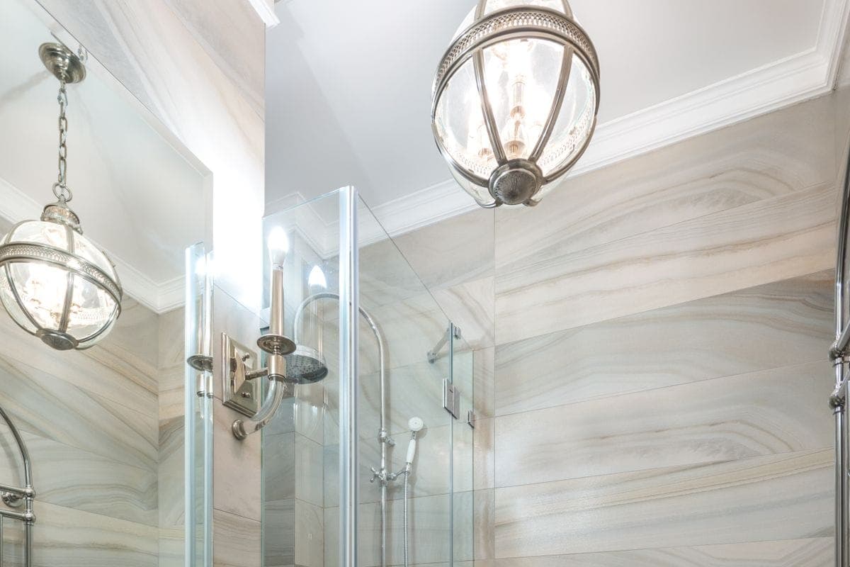 Baño alargado con paredes de azulejo veteado, lavabo sobre mueble blanco, mampara de ducha de cristal e inodoro al fondo