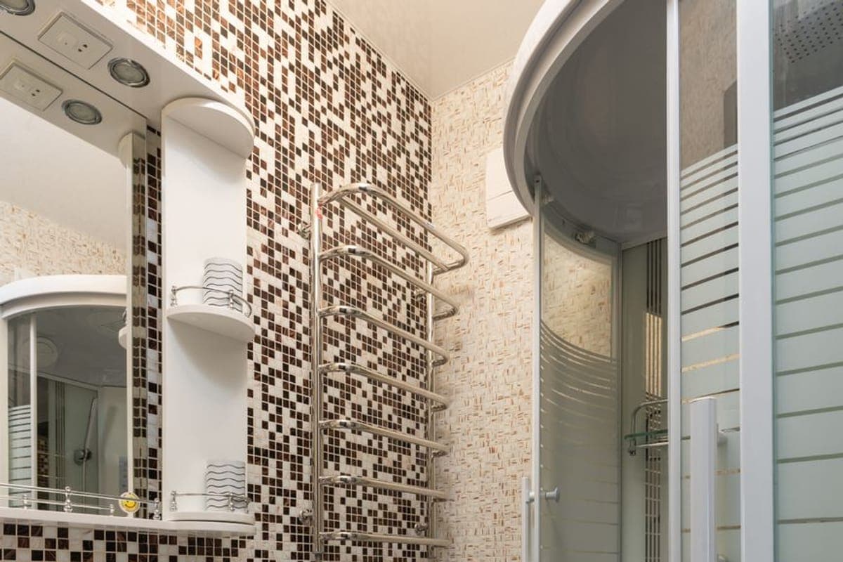 Baño estrecho de mosaico marrón con toallero radiador cromado, lavabo sobre mueble blanco y bañera con mampara curva