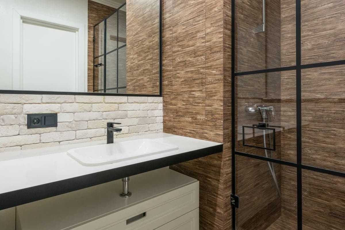 Baño con pared de azulejo imitación madera, lavabo sobre encimera oscura, zócalo de ladrillo visto con un enchufe y una mampara de ducha de perfil negro