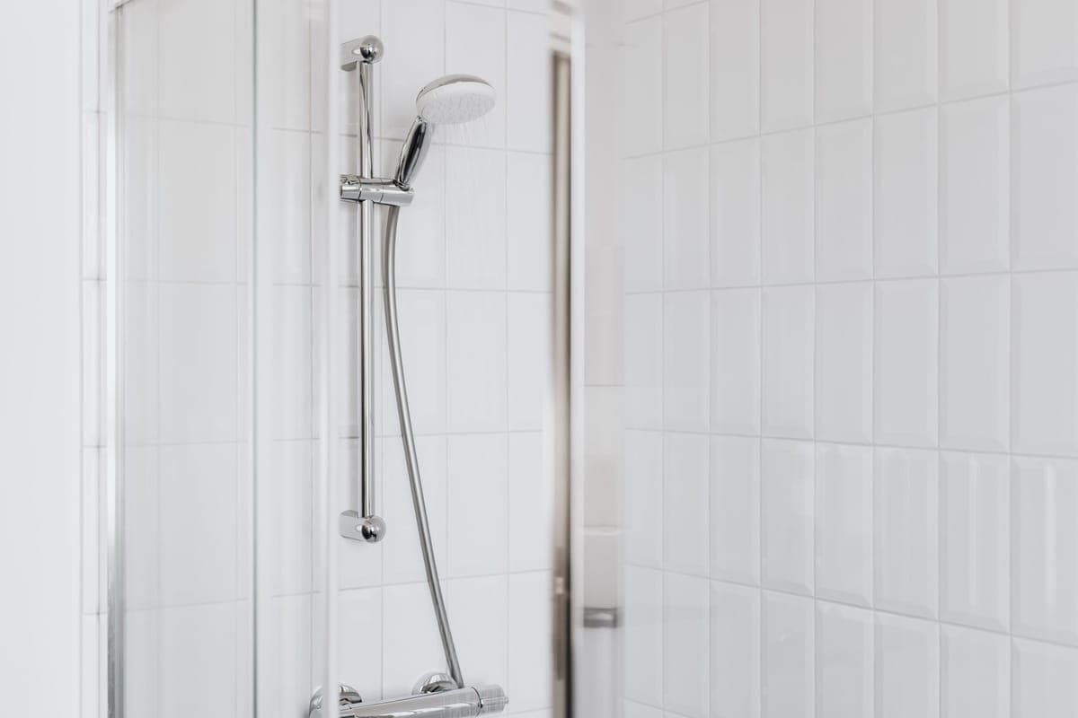 Barra de ducha cromada con teléfono y agua cayendo, en una ducha de azulejo blanco