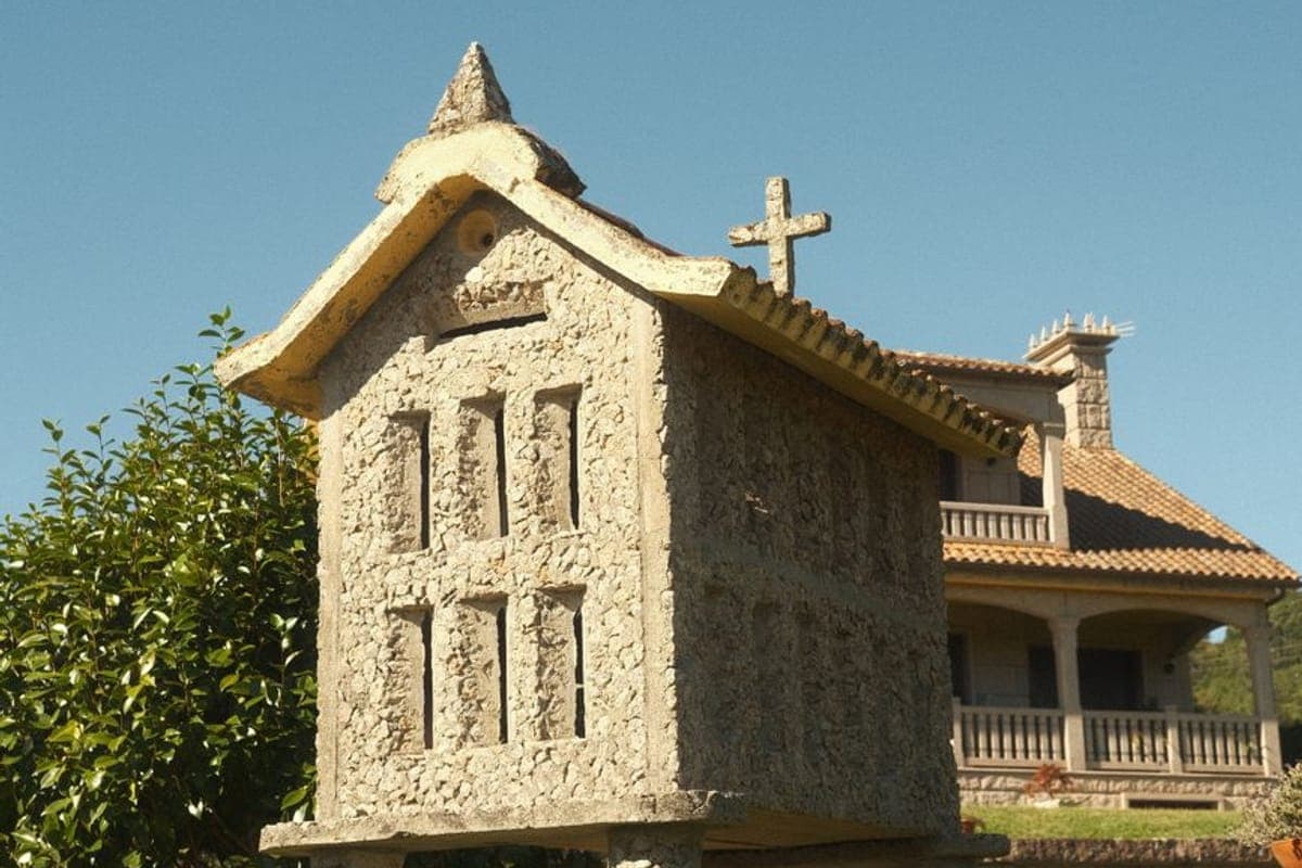 Hórreo de piedra con su cruz sobre un muro, y detrás una casa de galería con tejado de teja
