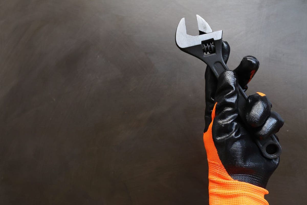Mano con guante negro y naranja de trabajo sujetando una llave ajustable sobre un fondo oscuro