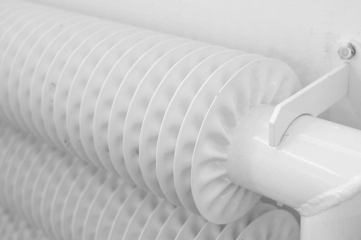 Radiador blanco de tubos aleteados visto muy de cerca, con la pared clara al fondo