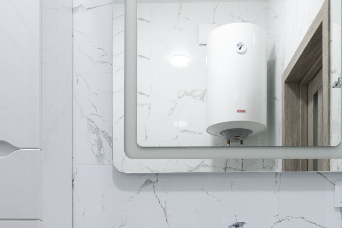 Lavabo blanco con grifo cromado bajo un espejo en el que se refleja un termo eléctrico vertical colgado en la pared de enfrente