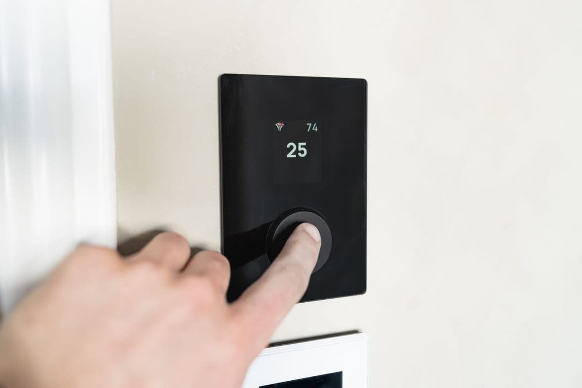 Un dedo pulsando la rueda de un termostato digital negro empotrado en la pared, con la pantalla marcando 25