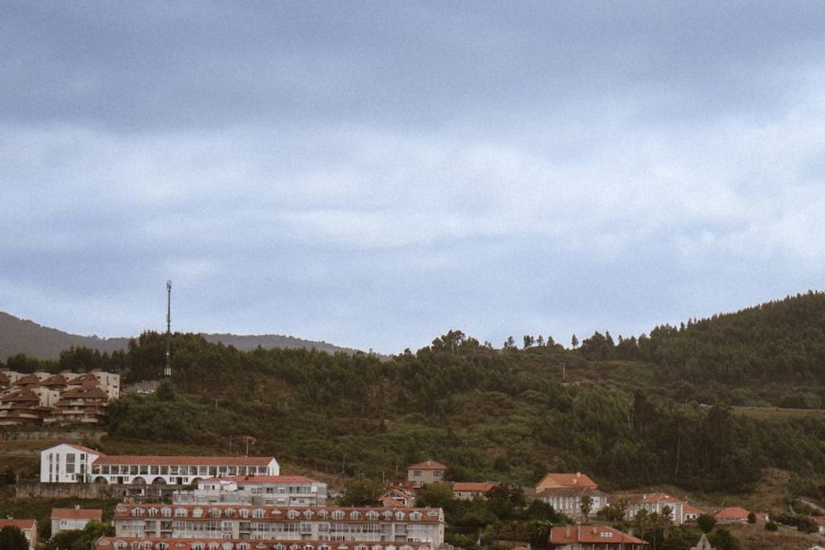 Villa costera de tejados rojos sobre una playa y una ría, con monte arbolado detrás y cielo nublado