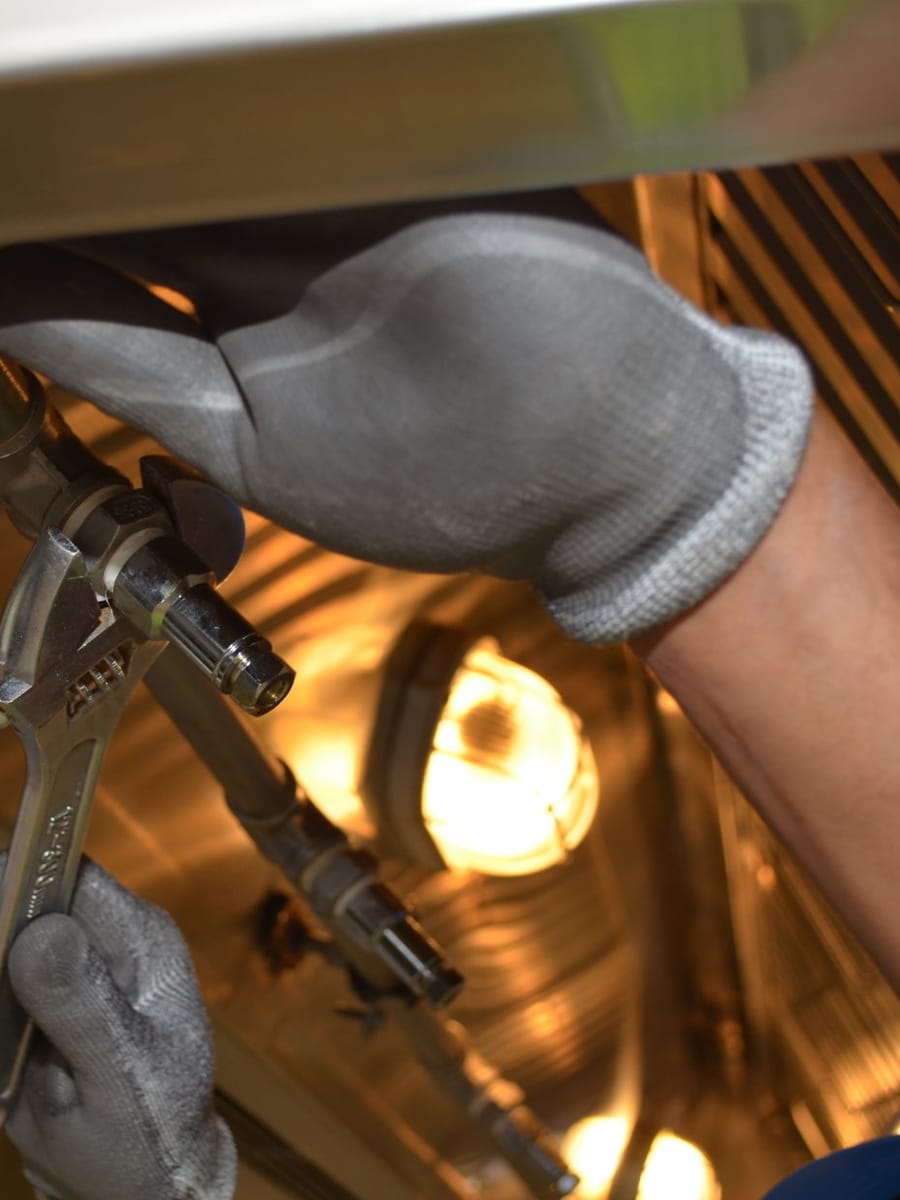 Dos manos con guantes grises aprietan con una llave ajustable un racor bajo un depósito de acero inoxidable