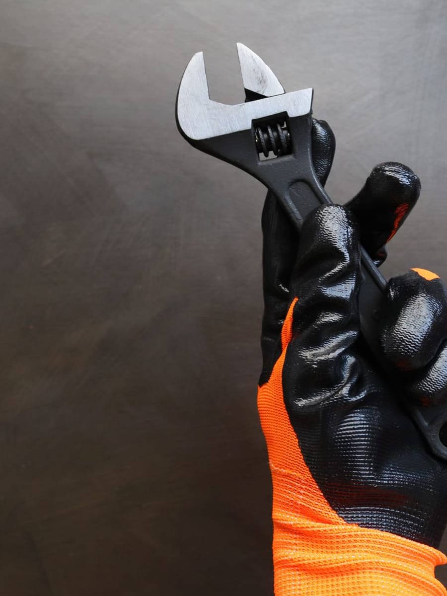Mano con guante negro y naranja de trabajo sujetando una llave ajustable sobre un fondo oscuro