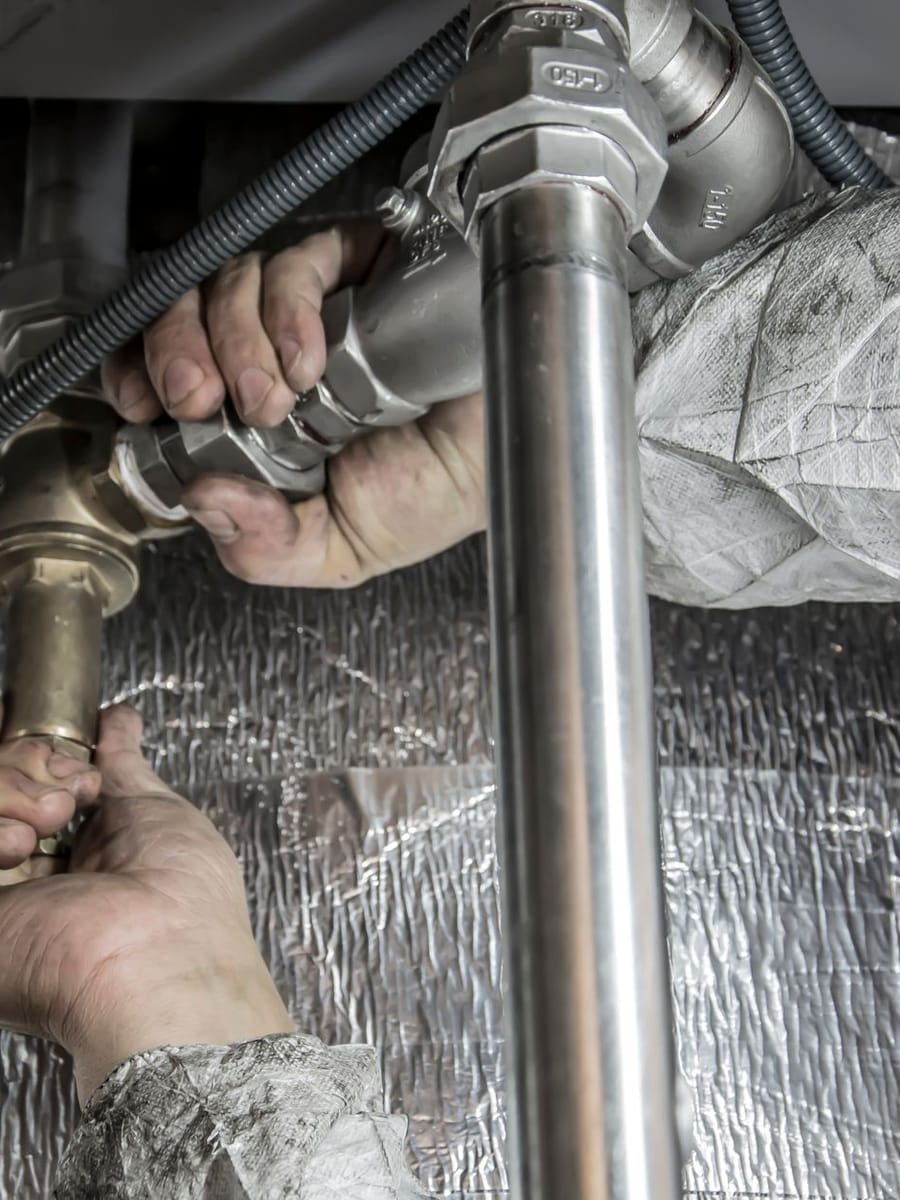 Dos manos aprietan un racor de latón bajo un aparato, entre tubos de acero y conductos forrados de aluminio