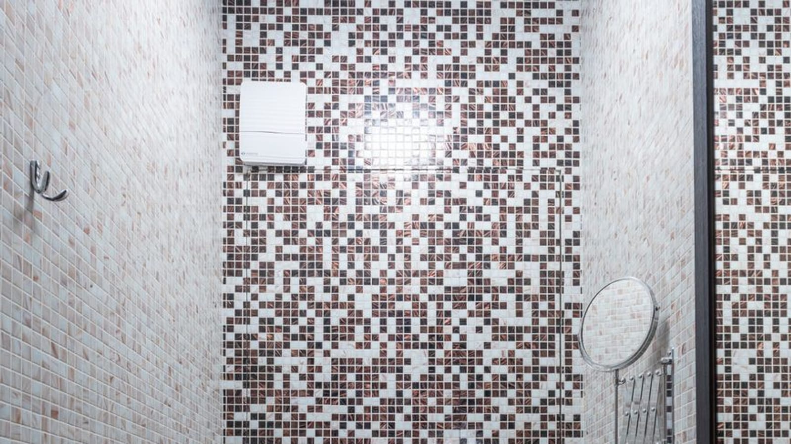 Aseo estrecho de mosaico marrón y blanco con inodoro suspendido, ducha de higiene, lavabo y extractor en la pared