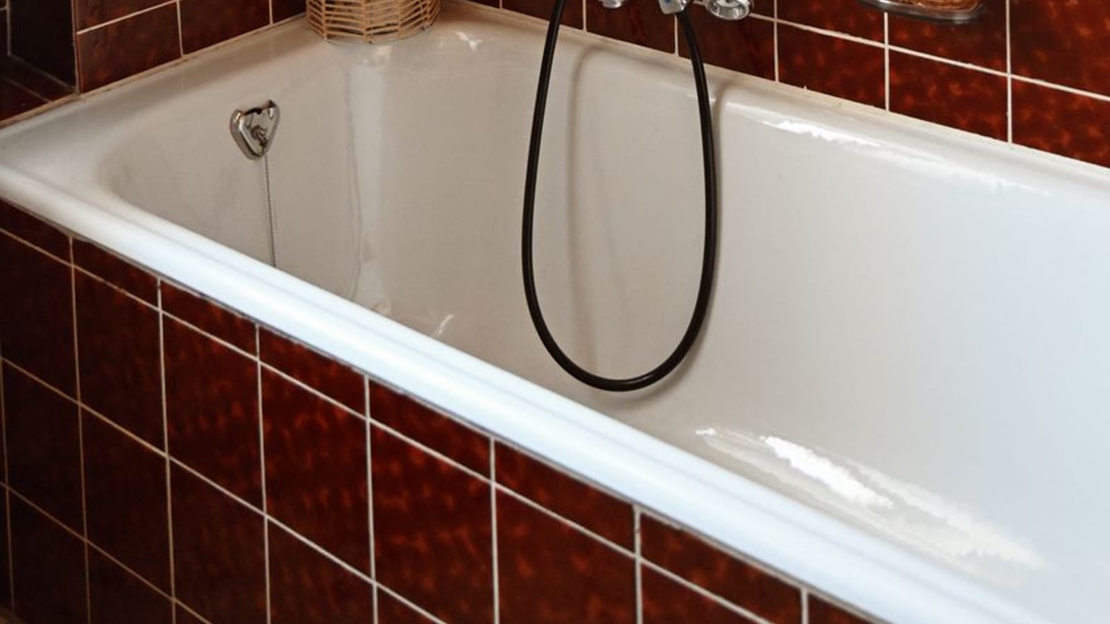 Bañera blanca con grifo cromado y flexible negro, en un baño alicatado de azulejo granate antiguo