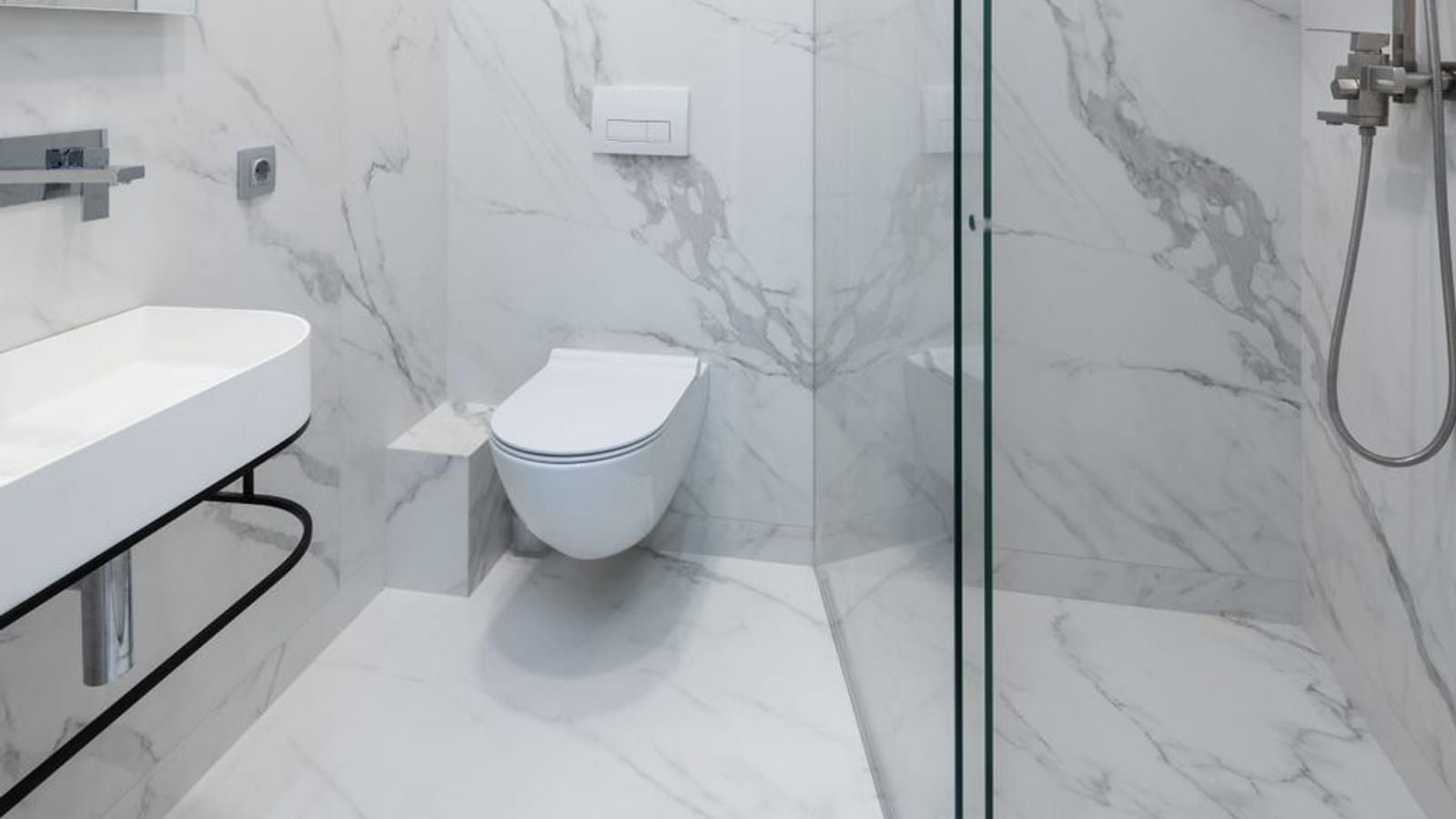 Baño de mármol blanco con inodoro suspendido y ducha de obra tras una mampara de cristal, con columna de ducha