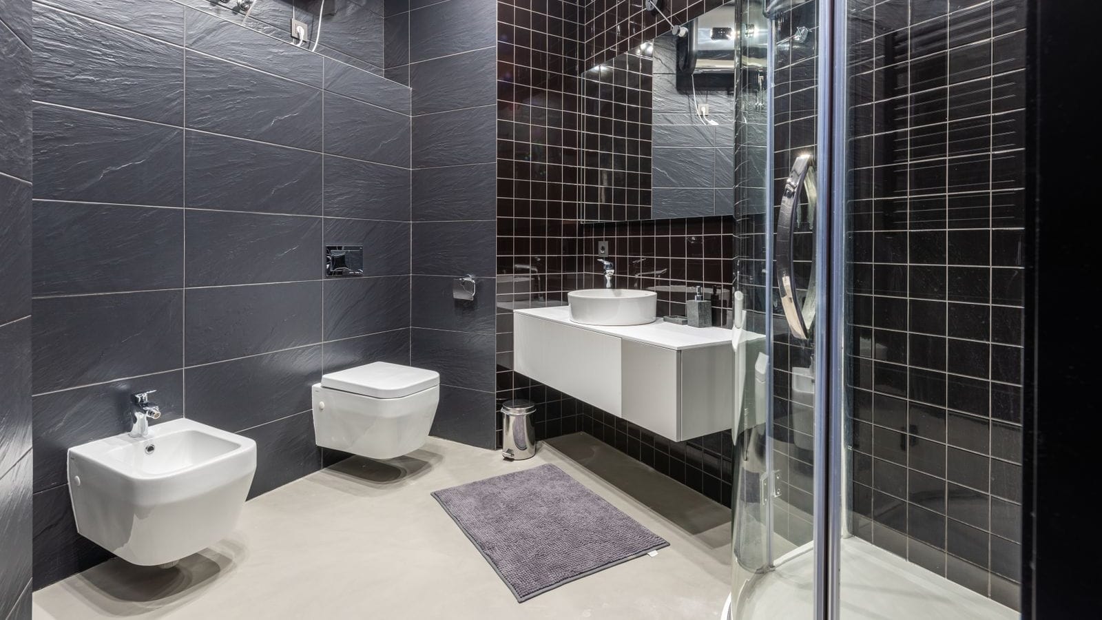 Baño de azulejo gris oscuro con un termo eléctrico horizontal colgado en lo alto de la pared, junto a la mampara de ducha y el lavabo suspendido