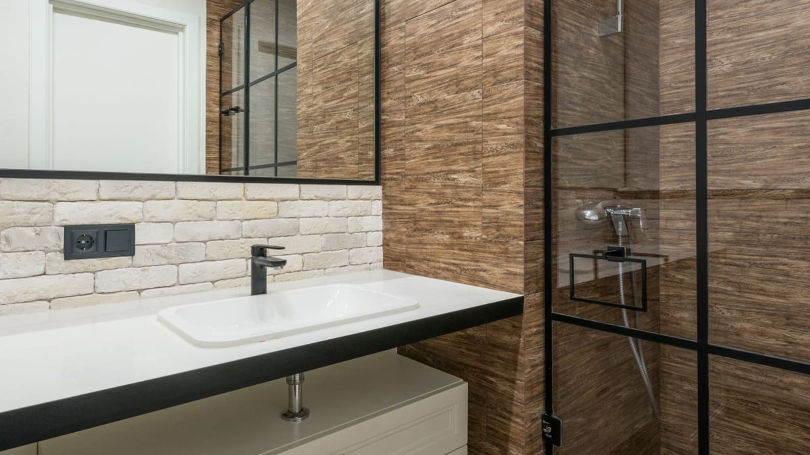 Baño con pared de azulejo imitación madera, lavabo sobre encimera oscura, zócalo de ladrillo visto con un enchufe y una mampara de ducha de perfil negro