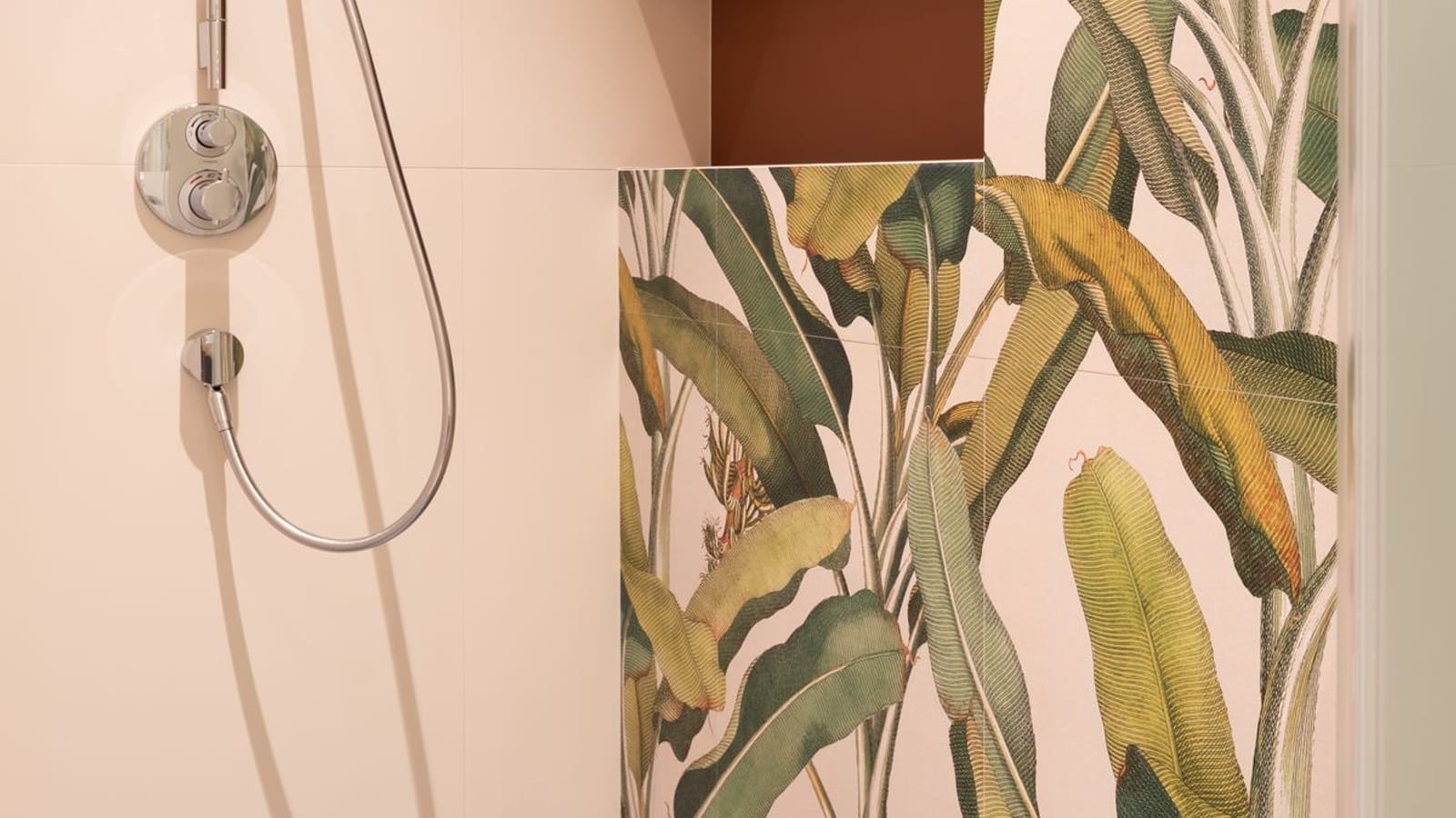 Rincón de ducha con grifo termostático cromado y flexible, junto a una pared alicatada con dibujo de hojas verdes y una hornacina