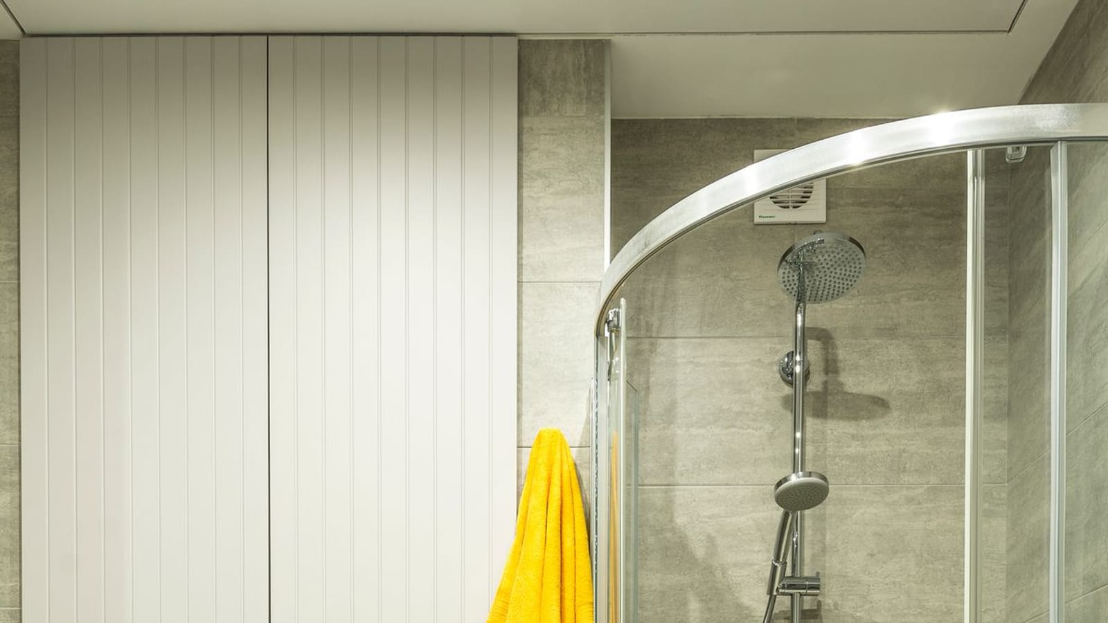Baño de azulejo gris con inodoro suspendido, armario blanco, una toalla amarilla colgada y una mampara de ducha de ángulo con columna cromada