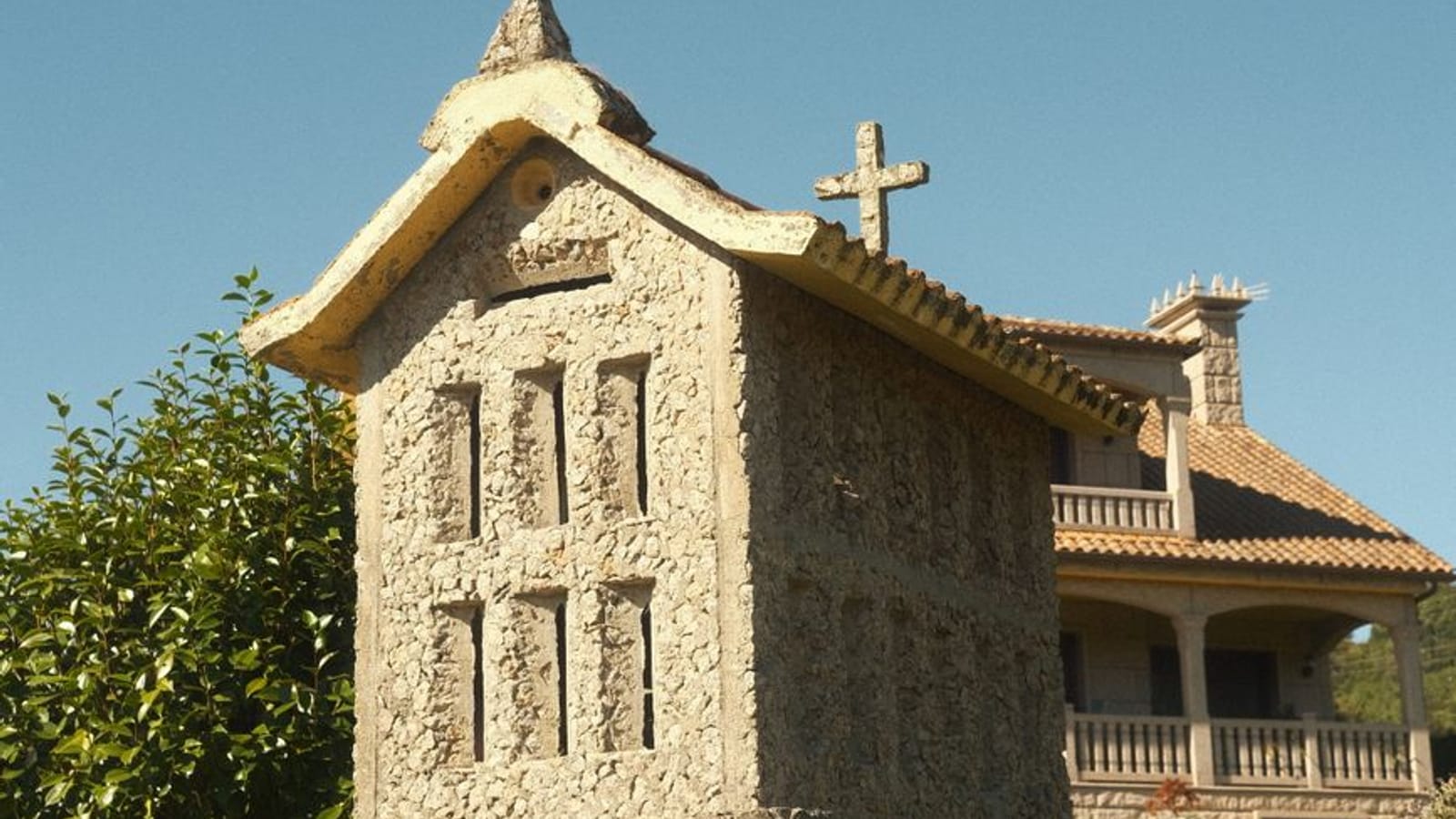 Hórreo de piedra con su cruz sobre un muro, y detrás una casa de galería con tejado de teja