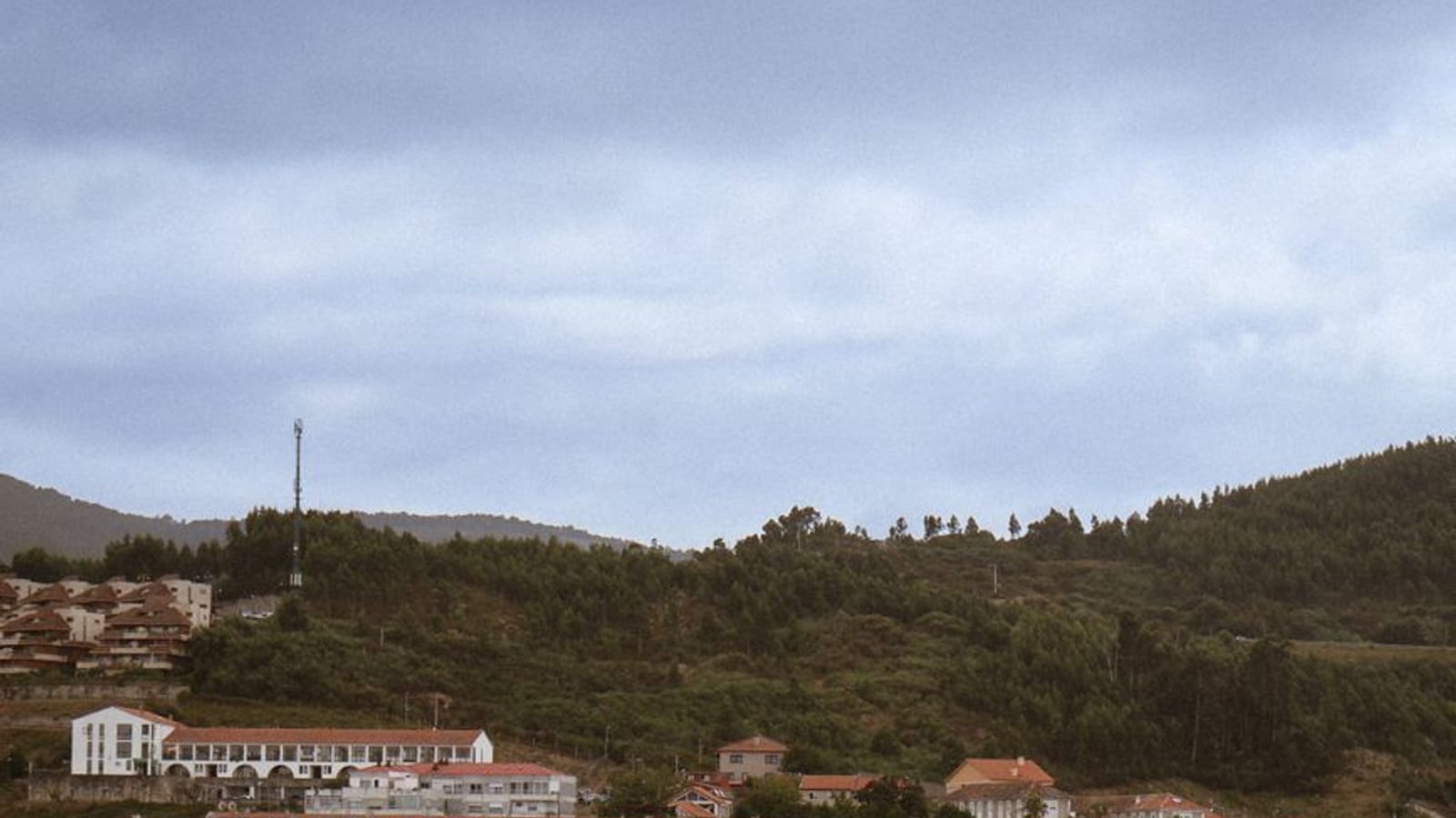 Villa costera de tejados rojos sobre una playa y una ría, con monte arbolado detrás y cielo nublado